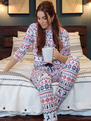 Woman wears the White Christmas Pyjamas whilst sitting on her bed with a hot drink.