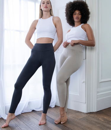 Two Model wearing the Everyday High Waisted Legging In colours Infinity Blue Marl and Oatmeal Beige. They are standing indoors, next to a white window and one model is leaning against a white wall.